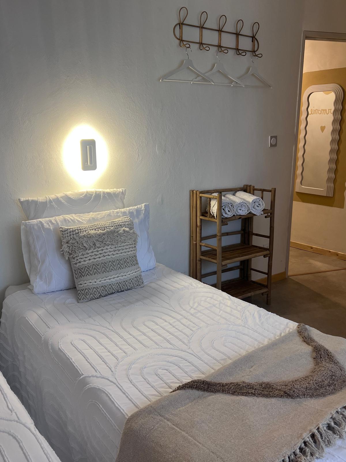 Chambre lumineuse aux matières naturelles avec lit simple, linge blanc, étagère en bambou et serviettes chez Origines be holistic.